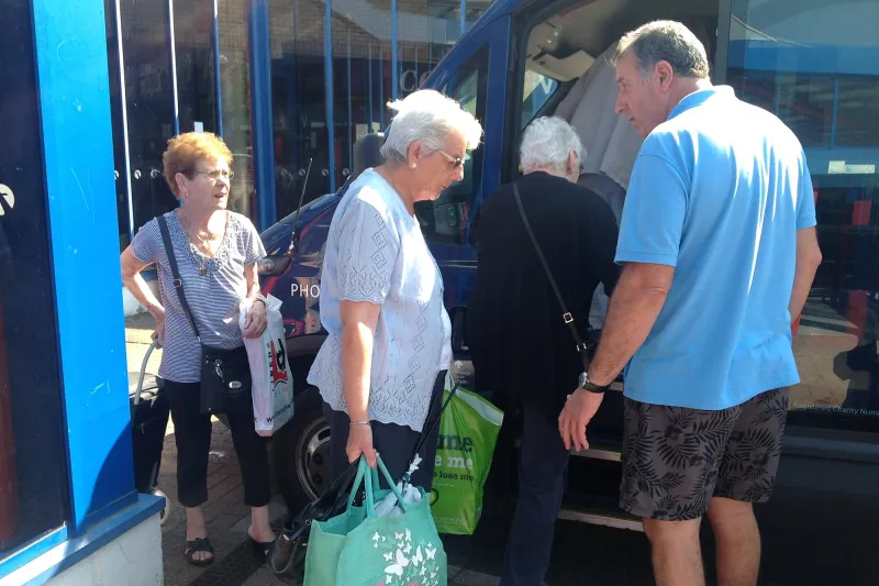 A driver helping passengers with their shopping bags onto a BCTS minibus outside the shops