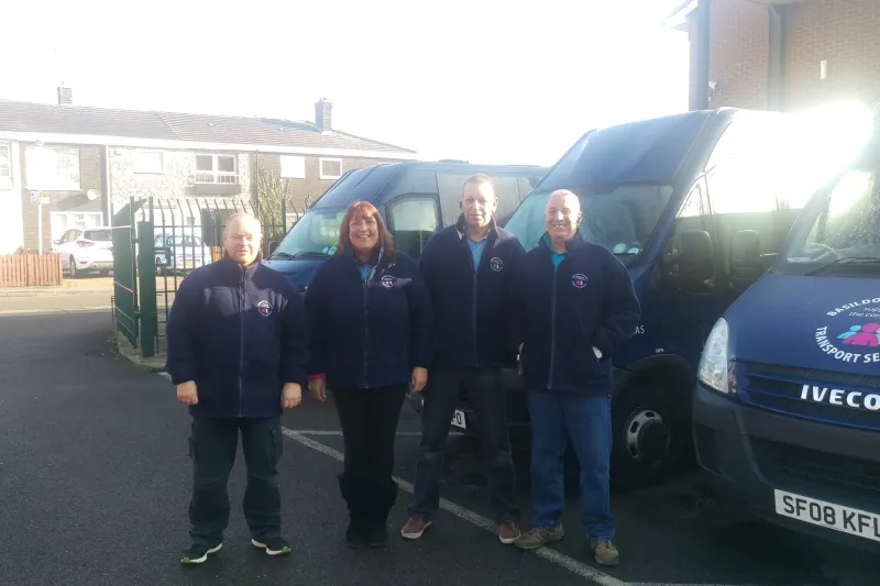 Four BCTS drivers in navy fleeces standing in front of two of our navy minibuses