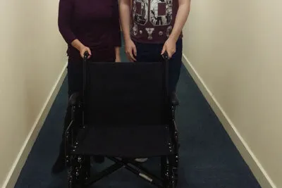 Two members of staff with a folding wheelchair ready to hire, in a corridor at the George Hurd Centre